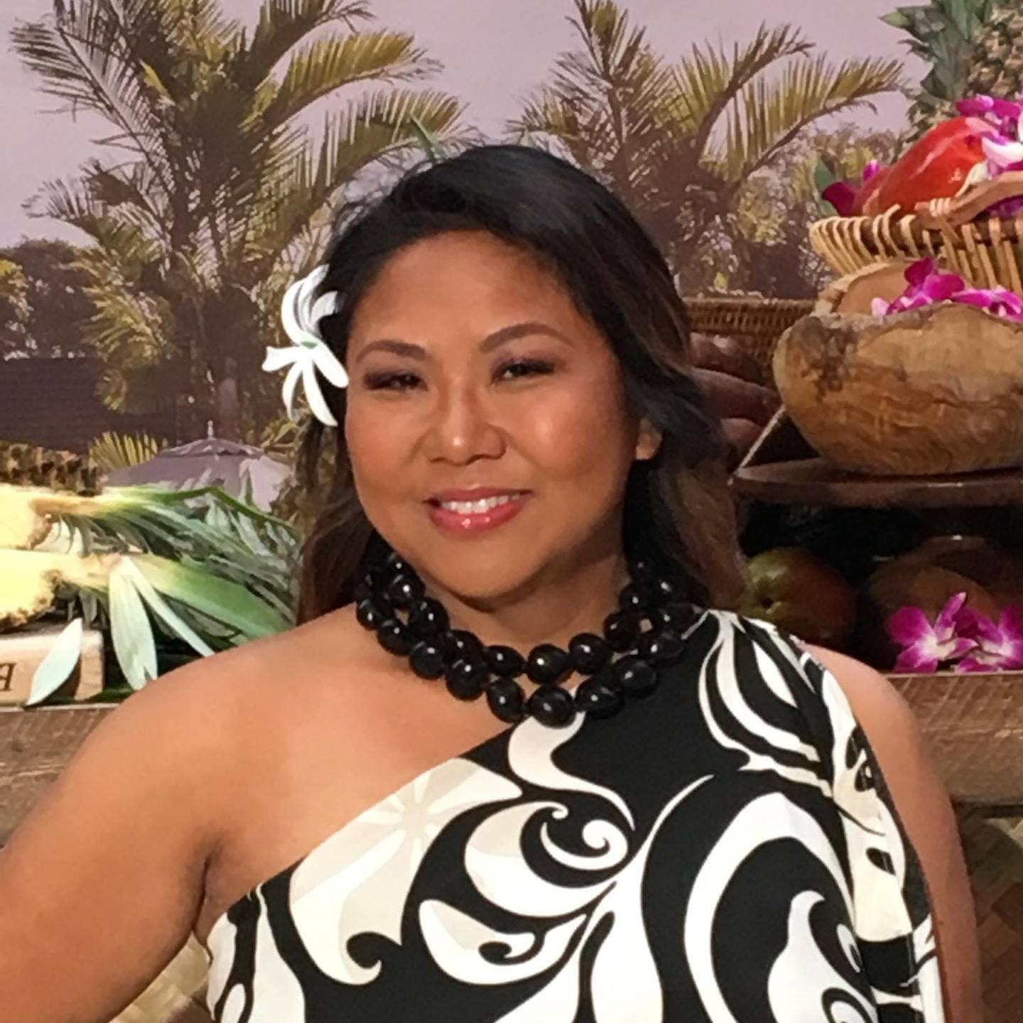 A background with color full flowers, with a light brown female in a black and white dress smiling. Behind her left ear, is a flower. 